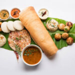 Group of South Indian food like Masala Dosa, Uttapam, Idli/idly, Wada/vada, sambar, appam, semolina halwa, upma served over banana leaf with colourful chutneys, selective focus